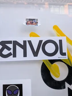 A small, roughly rectangular sticker with rounded top corners and a torn, irregular bottom edge. It features a black and white QR code structure, with a colorful, pixelated image embedded within its central data area. The embedded image displays a vibrant, multi-colored scene, possibly a crowd or a complex digital landscape, with prominent reds, blues, and whites against darker backgrounds, blending functional code with artistic expression.