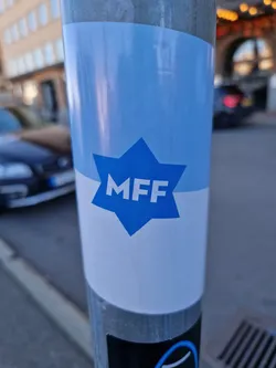 A blue and white sticker with the letters MFF in a star shape. The sticker is attached to a metal pole on a city street.