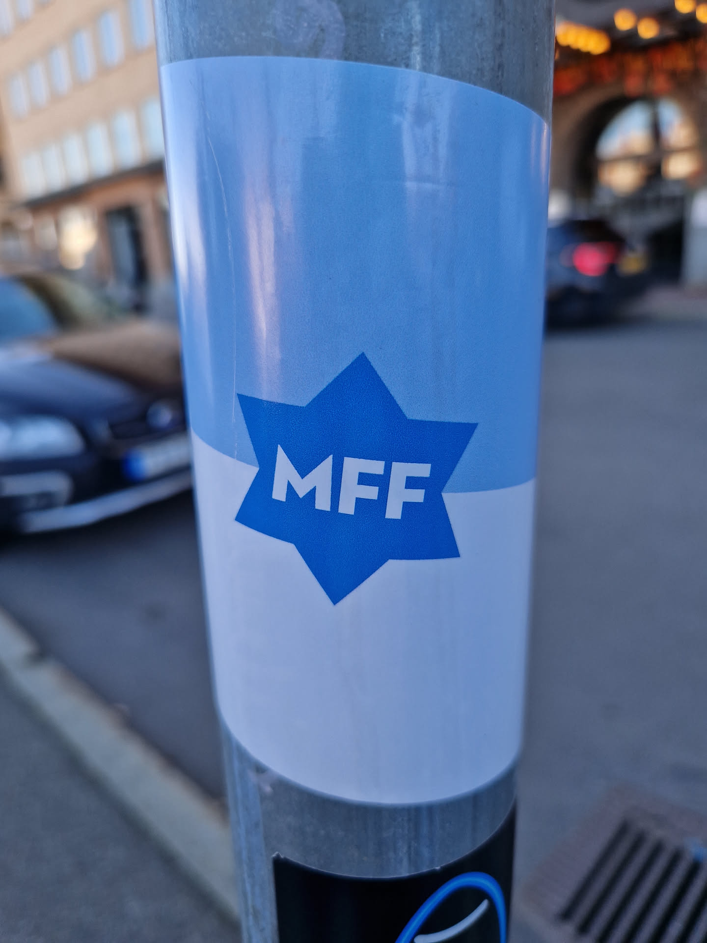 A blue and white sticker with the letters MFF in a star shape. The sticker is attached to a metal pole on a city street.