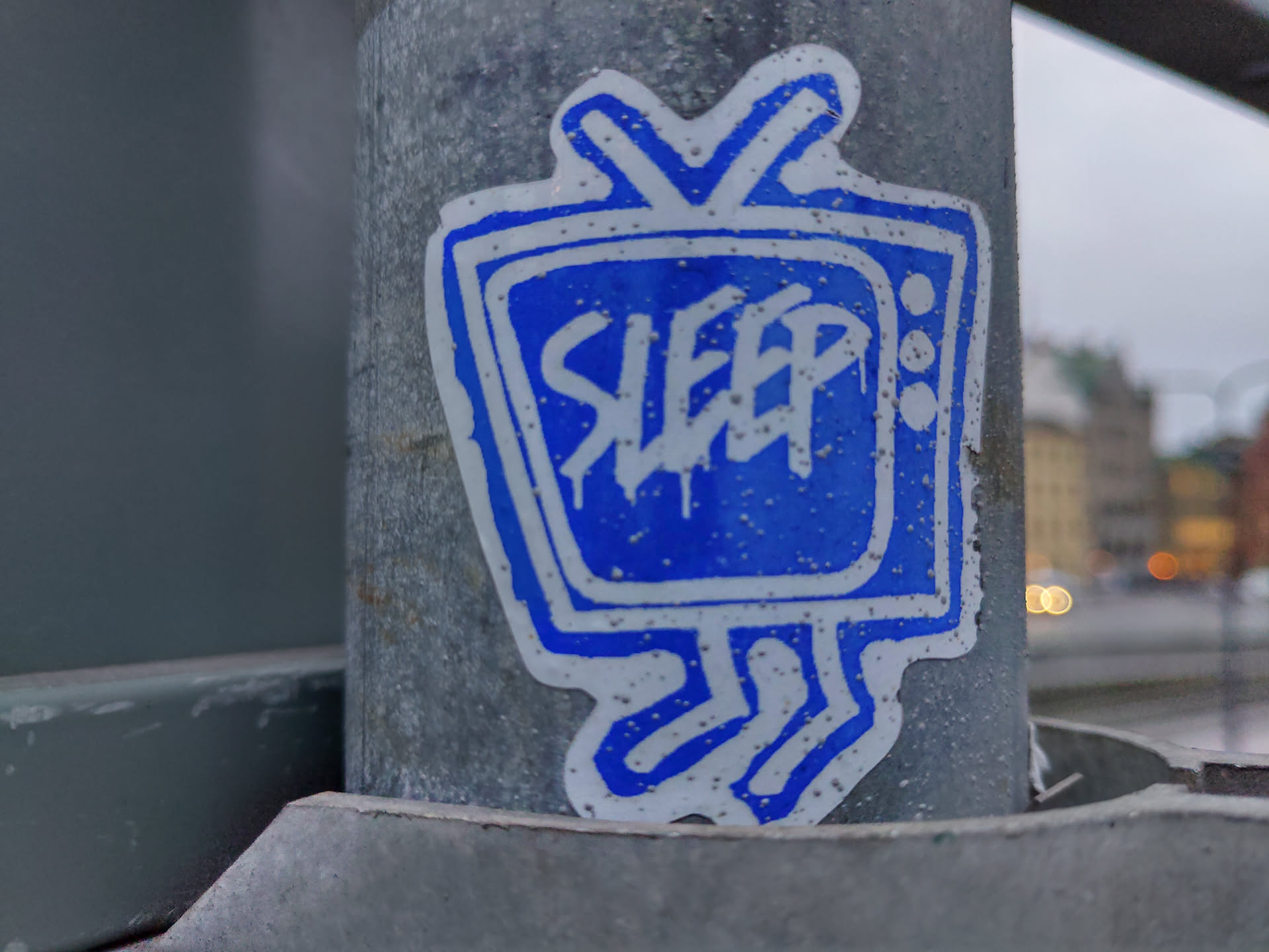 A blue sticker shaped like a retro TV with the word "SLEEP" written inside. It's attached to a metal pole outdoors. The style is simple and slightly grungy, with a hand-drawn feel.