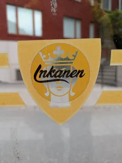 The sticker is shaped like a heraldic shield, primarily in a golden yellow color with a textured appearance, affixed to what appears to be a glass surface. In the center, there is a circular emblem featuring a simplified, stylized face in white/silver, topped by a detailed crown. The word "Inkanen" is written in a flowing, black script font across the center of the circle, partially overlapping the crown and the upper part of the face. The sticker shows signs of wear, with some cracking and scratches, suggesting age or exposure to elements. The background behind the glass is a blurred reflection of a red building and some foliage.