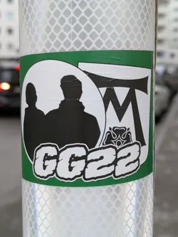 This is a white sticker with a green border featuring a silhouette of two figures, the letters 'MT', a stylized flower emblem, and the text 'GG22'.
