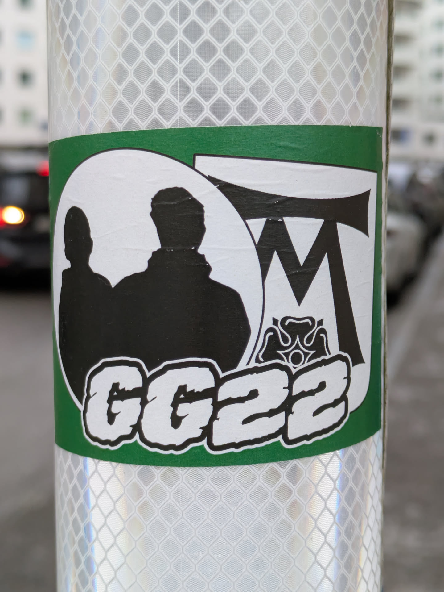 This is a white sticker with a green border featuring a silhouette of two figures, the letters 'MT', a stylized flower emblem, and the text 'GG22'.