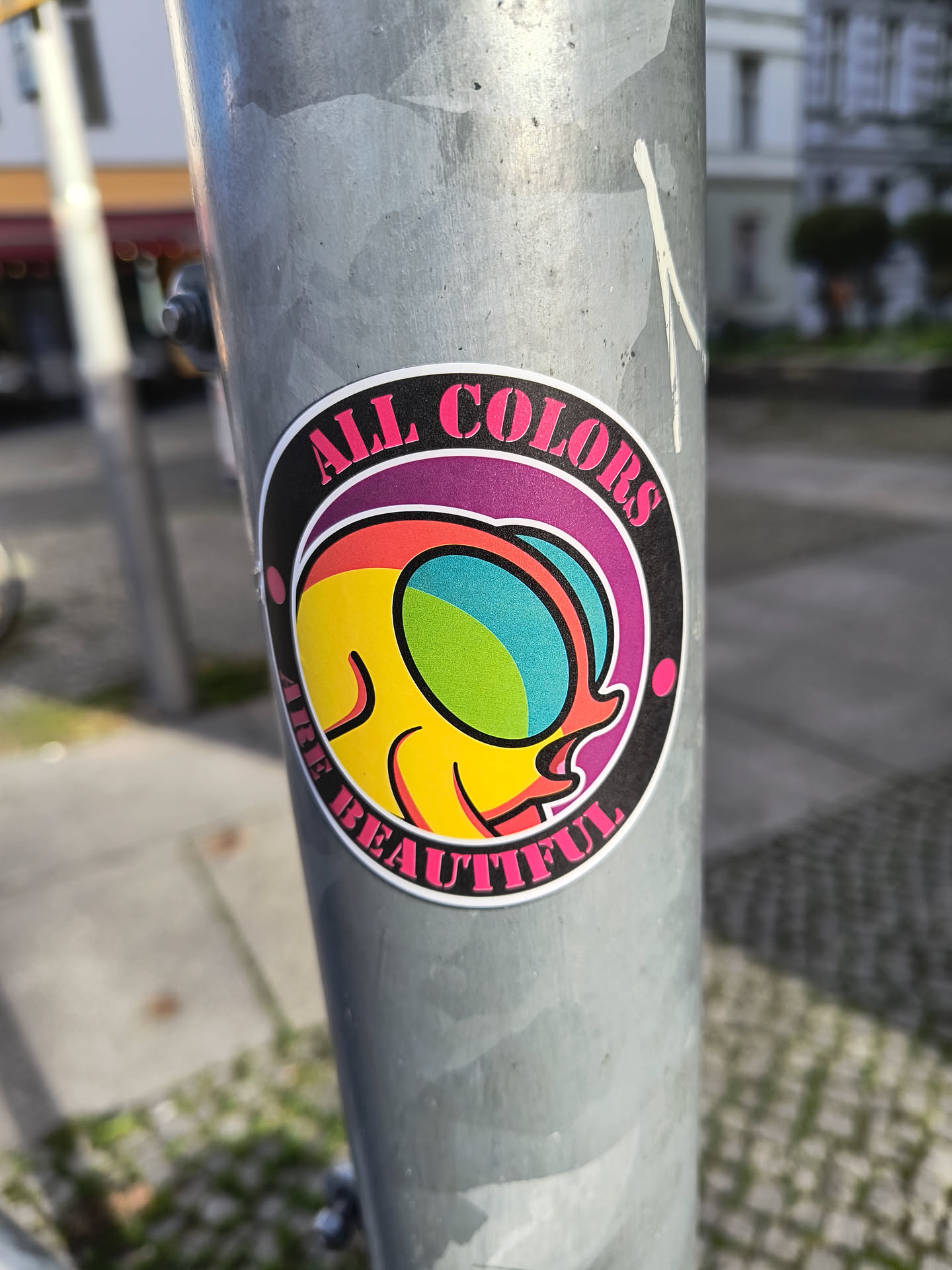 A round sticker with a colorful design and the text "All colors are beautiful". The design features an abstract figure with rainbow-like colors.