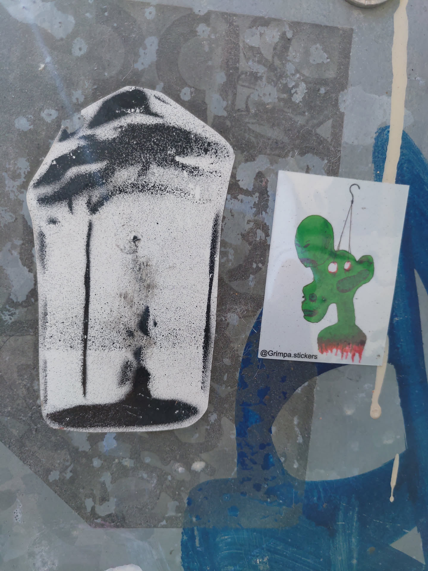 A sticker featuring a green alien-like figure hanging from a hook, with a dark drip or stain below it. The style is illustrative and slightly grotesque.