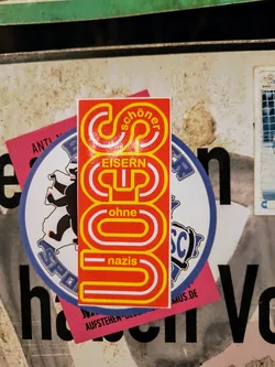 A sticker with a stylized design that says "Schöner Eisern Cup ohne Nazis" which translates to "nicer iron cup without Nazis". The sticker is red and yellow with white lettering. It's attached to a dirty surface with other stickers.