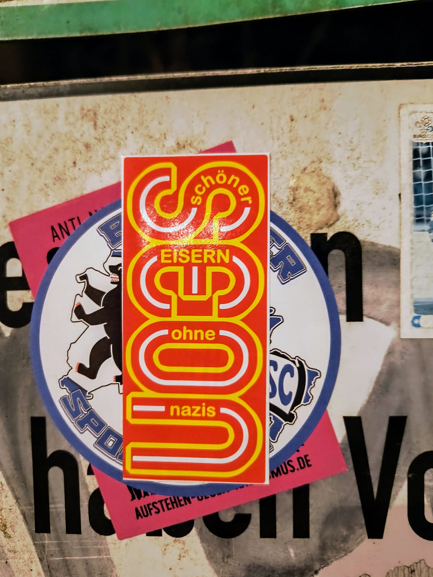 A sticker with a stylized design that says "Schöner Eisern Cup ohne Nazis" which translates to "nicer iron cup without Nazis". The sticker is red and yellow with white lettering. It's attached to a dirty surface with other stickers.