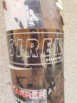 A sticker with the text "SIREN CREATIVES" in a sans-serif font with a white outline on a black background. The sticker is applied to a weathered surface with other stickers and graffiti.
