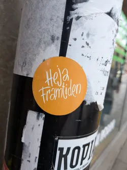 The sticker is perfectly circular with a bright, solid orange background. Centered on the sticker is the phrase 'Heja framtiden' written in a flowing, handwritten-style script font in white. The letters have a friendly, informal appearance. The surface of the sticker shows minor imperfections, possibly small water droplets or dust, and a slight sheen from ambient light. It is adhered to a textured, multi-layered pole covered with other torn paper and black sections.