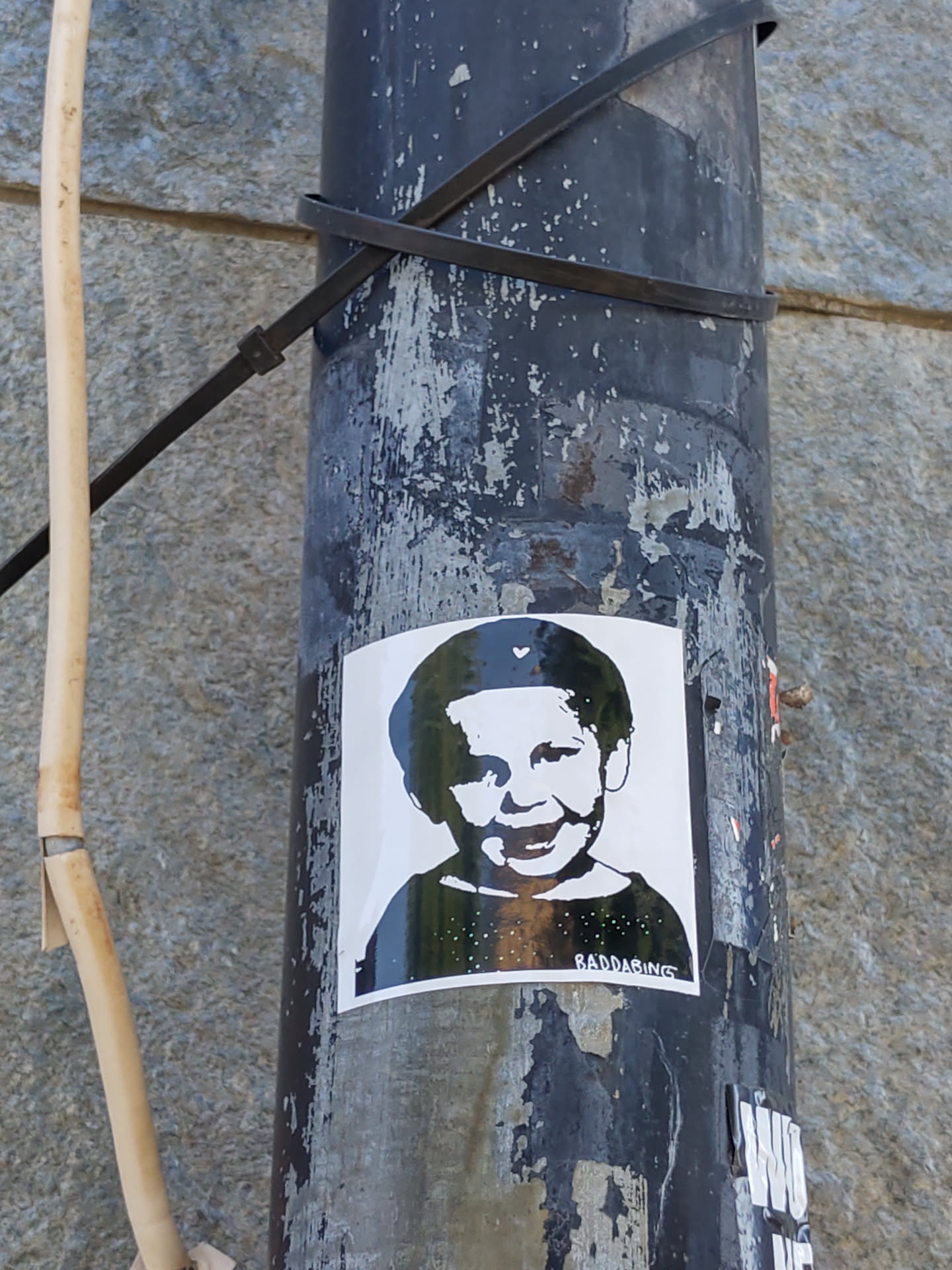 A sticker featuring a black and white stencil of a smiling child. The sticker has a minimalist design and is affixed to a weathered pole. The artist's tag, raddaR_nyc, is visible at the bottom.