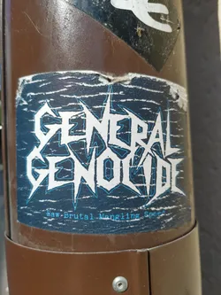A worn sticker with the band name General Genocide and the tagline "Raw Brutal Mangling Speed". The sticker has a dark blue background with white and light blue text. It appears to be stuck on a brown metal pole.