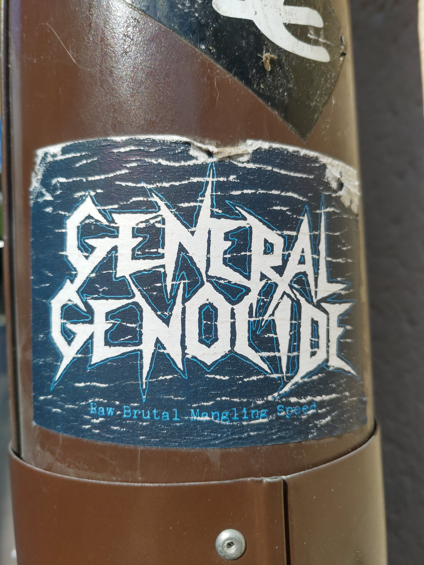 A worn sticker with the band name General Genocide and the tagline "Raw Brutal Mangling Speed". The sticker has a dark blue background with white and light blue text. It appears to be stuck on a brown metal pole.