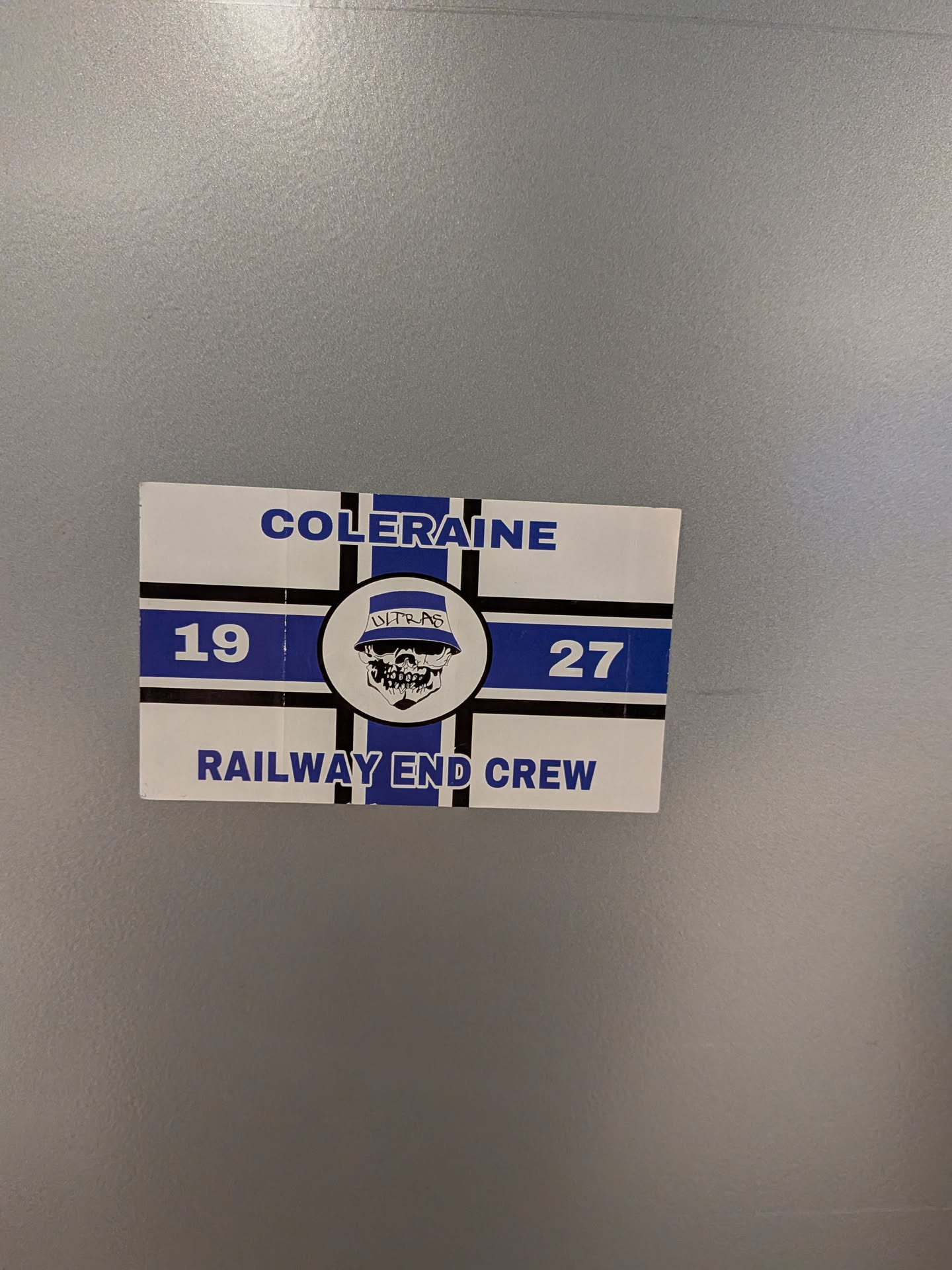 A rectangular sticker with a white background, featuring a prominent blue and black design. At the top, 'COLERAINE' is written in bold blue capital letters. Below this, a wide blue horizontal stripe is intersected by a black vertical stripe, forming a cross-like pattern. On the left side of the blue stripe, the number '19' is in white, and on the right, '27' is in white. Centered within the cross is a circular emblem depicting a stylized white skull with black outlines and details, wearing a blue bucket hat that has 'ULTRAS' written across it in white. Below the main design, 'RAILWAY END CREW' is written in bold blue capital letters. The sticker has slightly worn edges and is affixed to a textured grey surface.