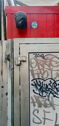 A pixel art sticker with blue, green, yellow, and purple pixels is stuck to a red metal wall, right beside a black security camera. The sticker is small and square, displaying an abstract, colorful design. Below the camera and wall, there is a white door with graffiti.