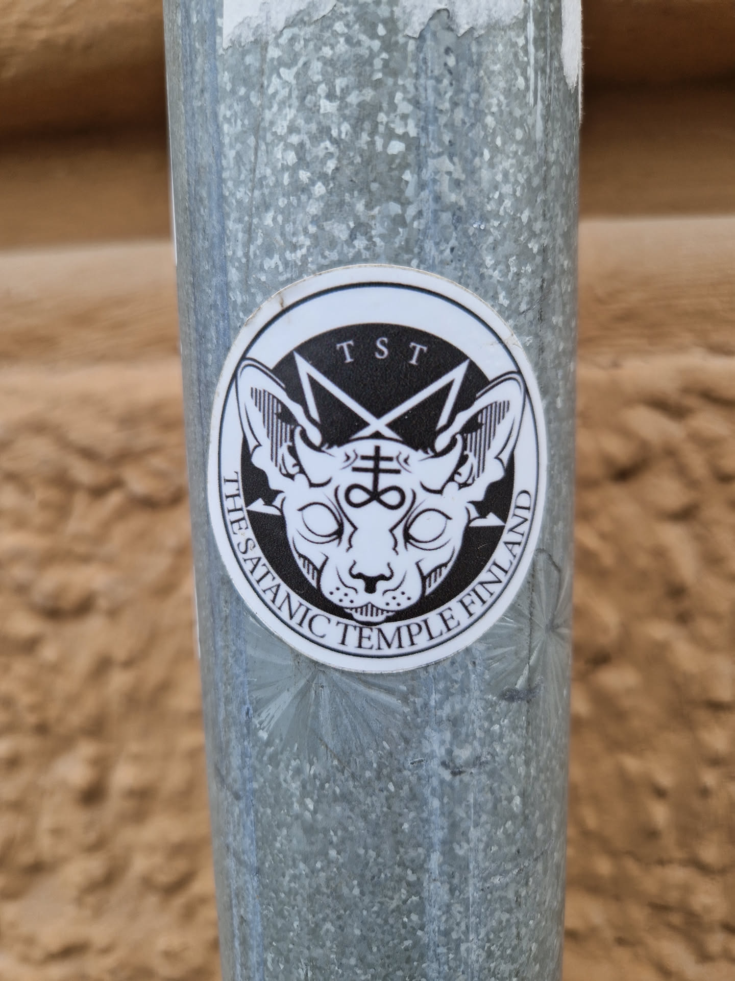 A round sticker with a black and white illustration of a sphinx-like cat's head with bat-like ears. The head is encircled by a pentagram and the text "TST" appears at the top. At the bottom it says "The Satanic Temple Finland".