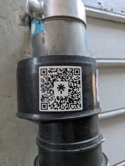 A white square sticker featuring a black QR code is prominently displayed on a dull black pipe, which is part of a larger plumbing or conduit system. The QR code contains a large, stylized asterisk (*) symbol in its center, adding a unique visual element to the standard code pattern. The pipe is connected to a larger metallic pipe assembly above it, which is secured by a metal clamp. The background shows a light-colored building surface, possibly stucco or siding.