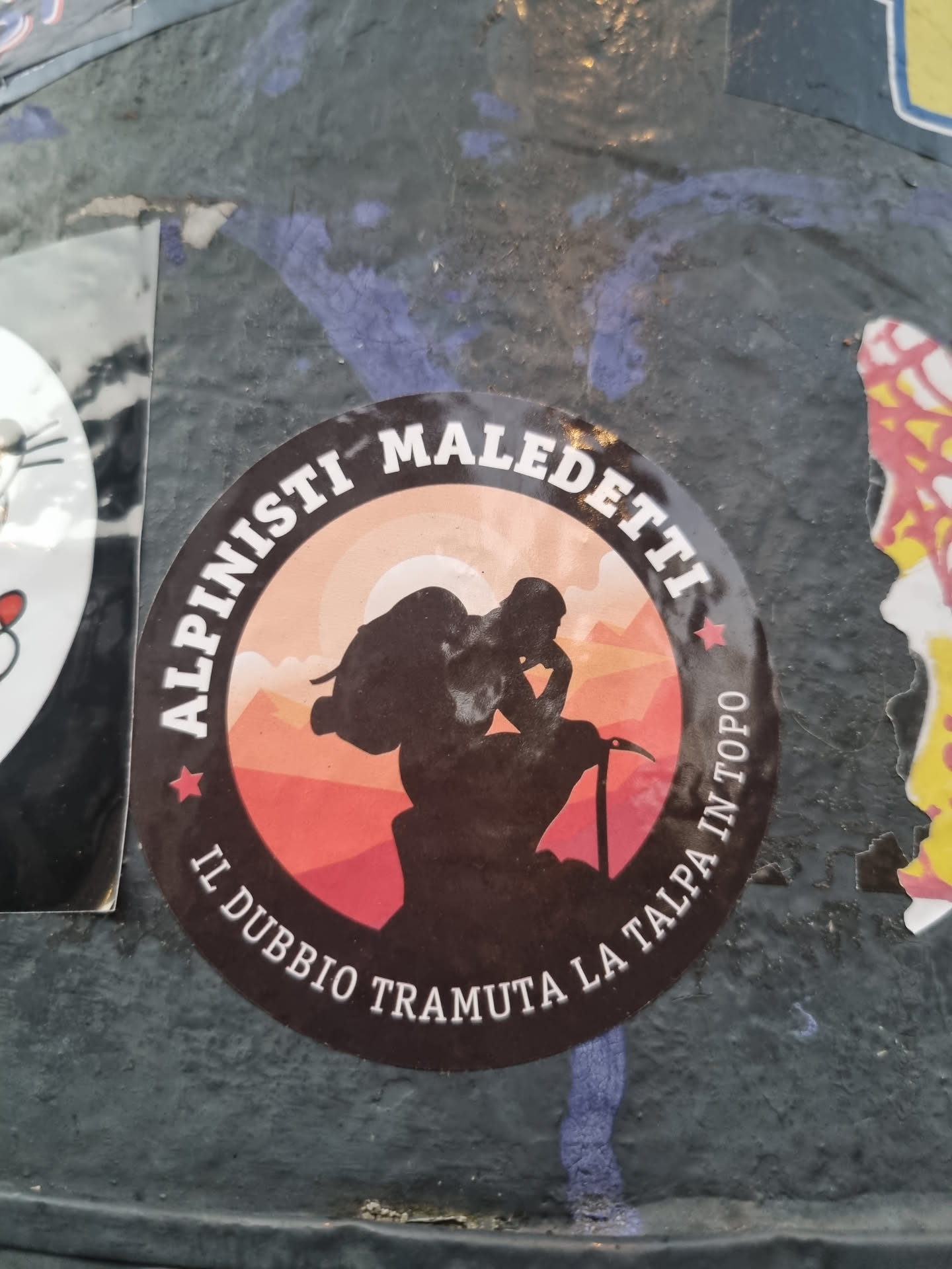 Round sticker with a dark background, depicting the silhouette of a climber against a sunset backdrop of mountains. The text around the circle reads "Alpinisti Maledetti" at the top, and "Il dubbio tramuta la talpa in topo" at the bottom. The overall design suggests a theme of mountaineering or climbing, with a slightly edgy or rebellious tone.