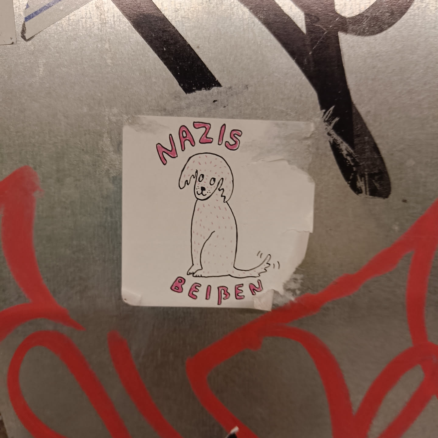 A sticker with a drawing of a dog and the text "Nazis beißen" which translates to "Nazis bite". The sticker is placed on a wall with other graffiti.