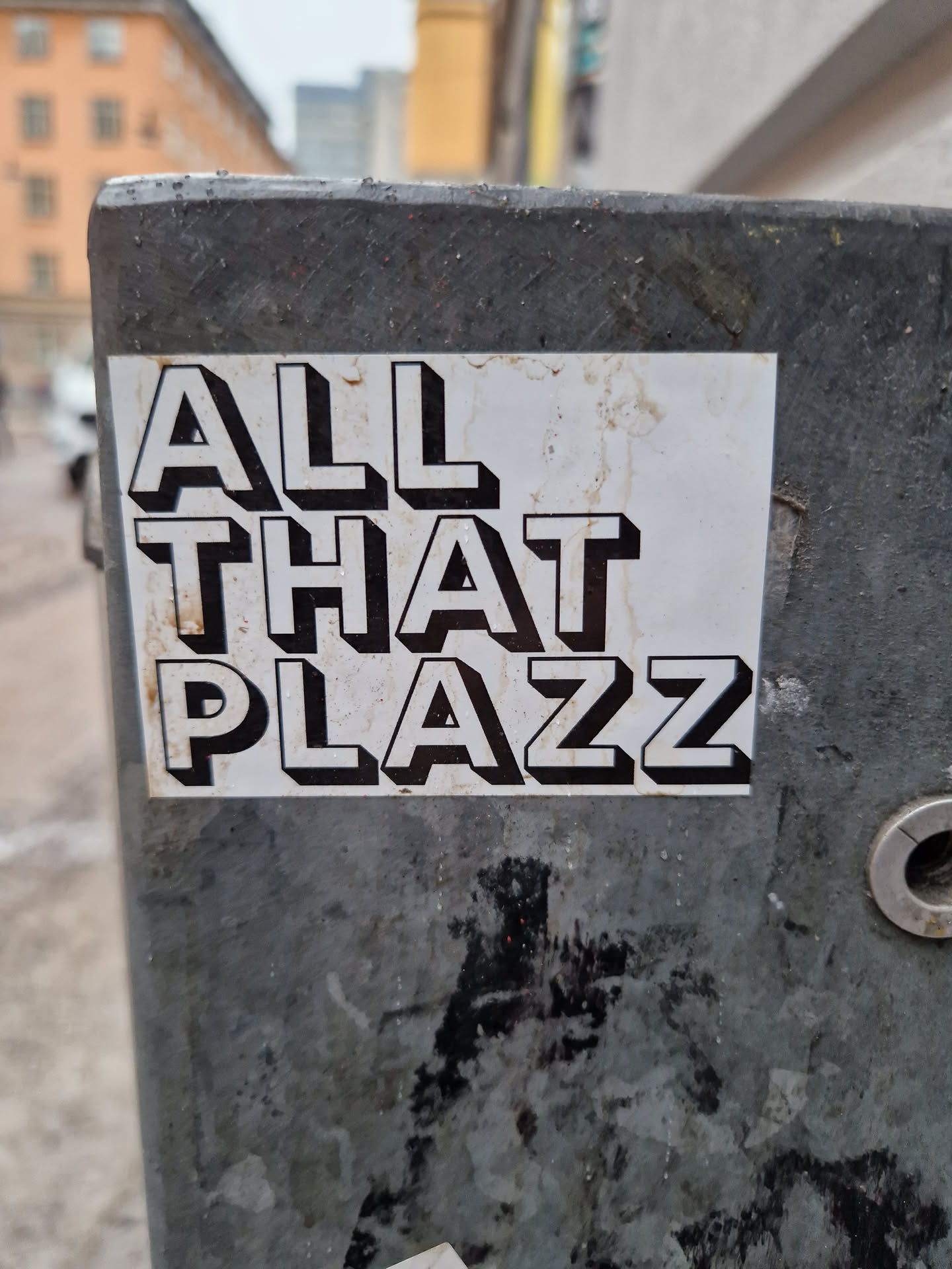 A sticker with the text "ALL THAT PLAZZ" in a bold, three-dimensional style font. The sticker is affixed to a weathered gray metal surface.