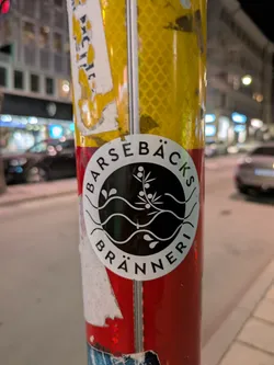 A round black and white sticker with the text 'Barsebäcks Bränneri' around the edge of the circle. The center of the sticker contains an image of waves of water and a plant with berries.