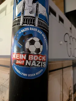 Street sticker An oval sticker adhered to a black cylindrical post. It features a bright blue circular background showing a classic black-and-white soccer ball smashing a black swastika, causing it to break apart. Across the upper edge, the slogan reads '» NAZIS RAUS AUS DEN STADIEN! «' in white capital letters. Across the center is a red-and-white block logo reading 'KEIN BOCK auf NAZIS', and the bottom curve reads '• FUSSBALLFANS GEGEN RECHTS •'.