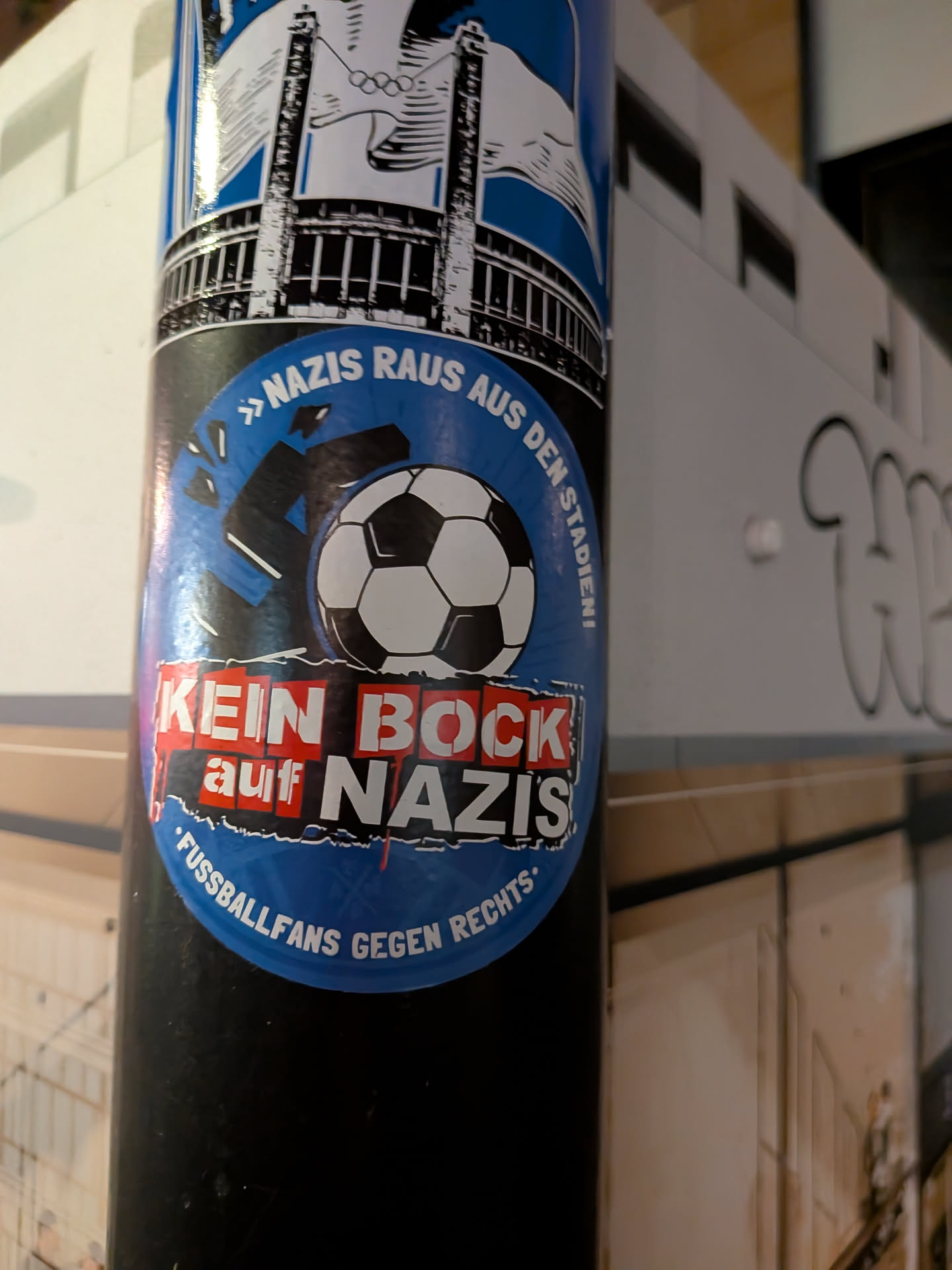 An oval sticker adhered to a black cylindrical post. It features a bright blue circular background showing a classic black-and-white soccer ball smashing a black swastika, causing it to break apart. Across the upper edge, the slogan reads '» NAZIS RAUS AUS DEN STADIEN! «' in white capital letters. Across the center is a red-and-white block logo reading 'KEIN BOCK auf NAZIS', and the bottom curve reads '• FUSSBALLFANS GEGEN RECHTS •'.