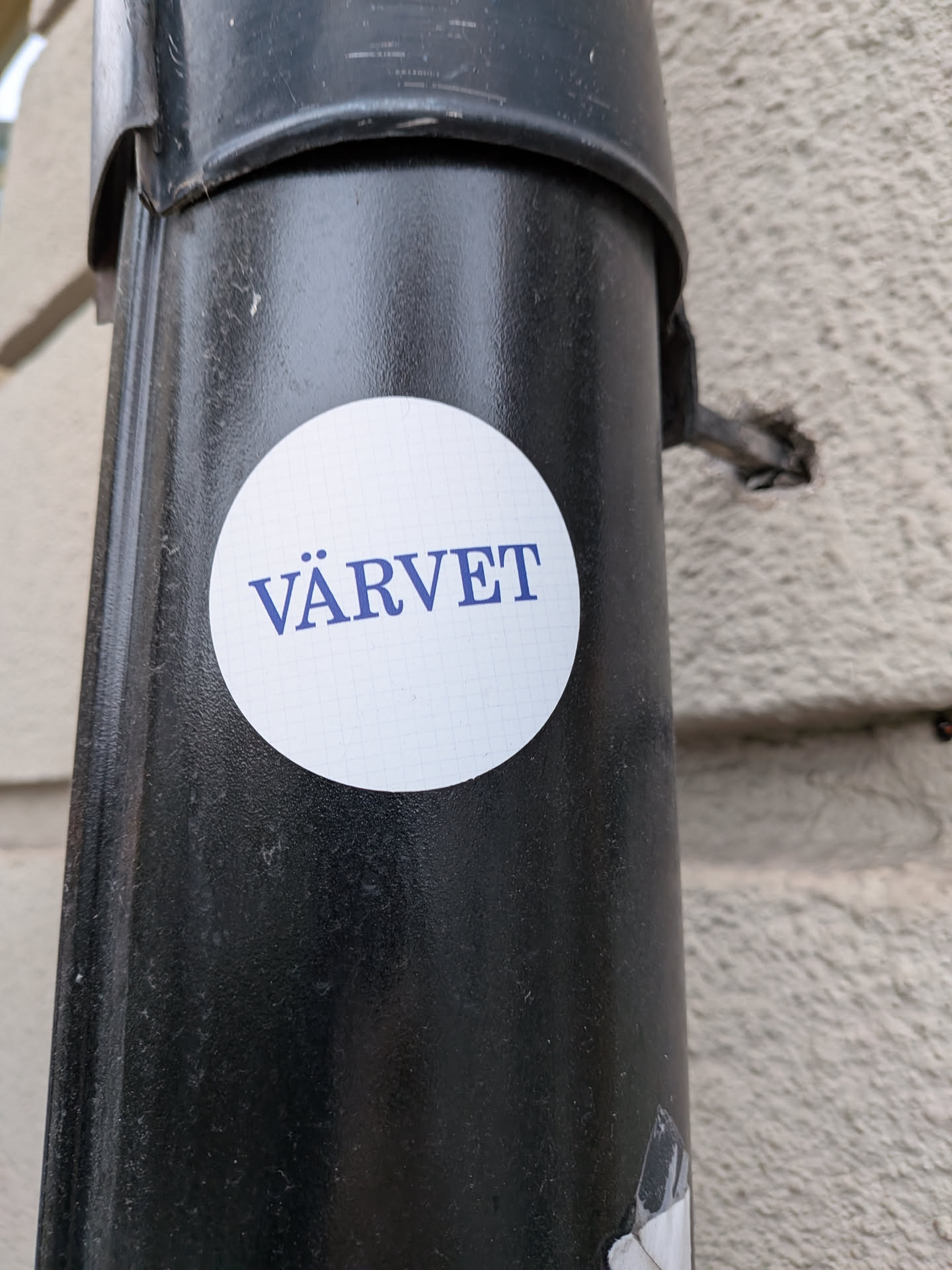 A round sticker adhered to a black metal drainpipe. The sticker features a white background with a subtle light grid pattern, reminiscent of graph paper. Centered across the sticker is the Swedish word 'VÄRVET' printed in a bold, dark blue serif font.