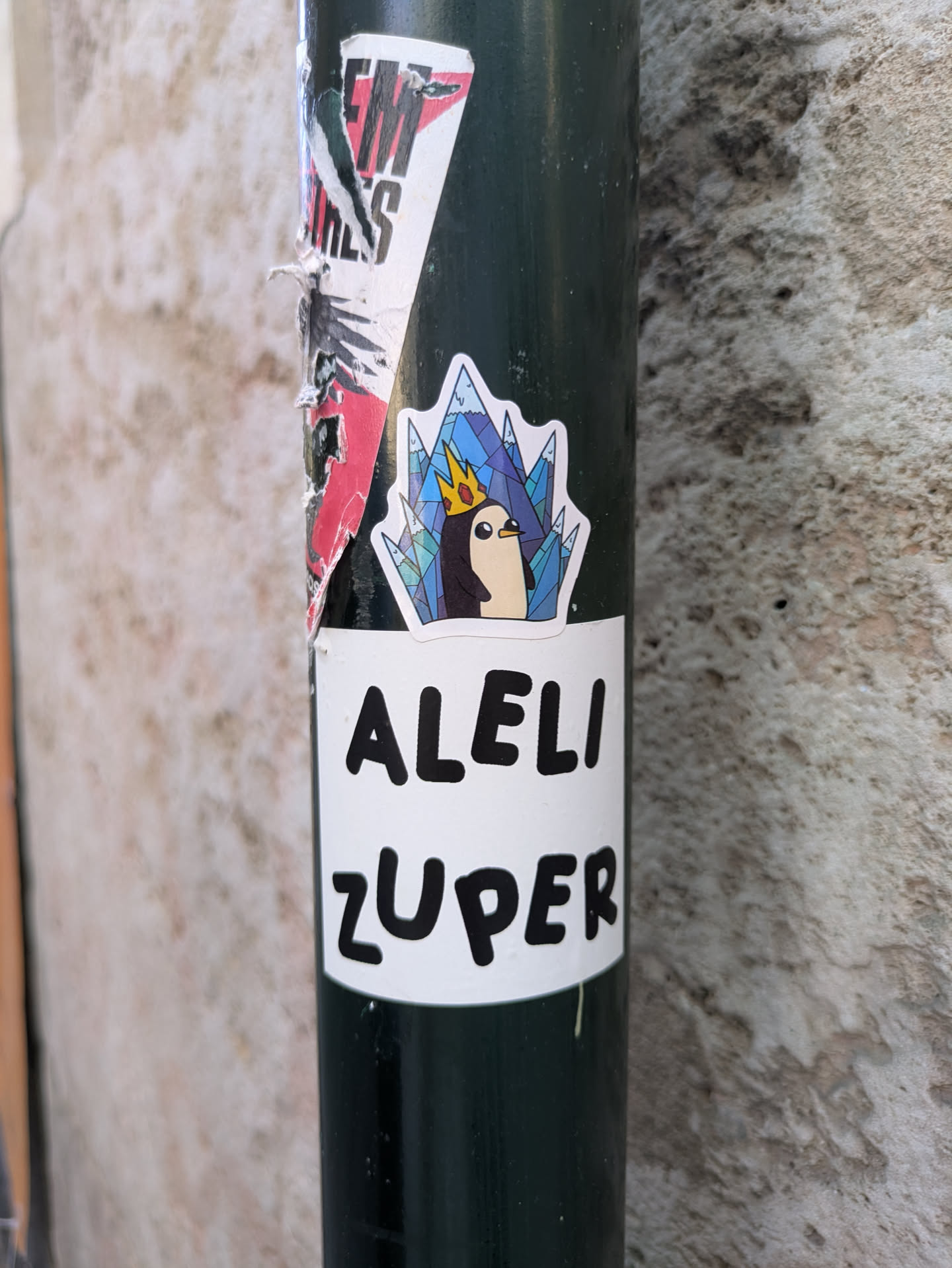 A sticker on a pole features Gunter the penguin from Adventure Time wearing the Ice King's crown in front of an icy mountain range, with the text "ALELI ZUPER" below.