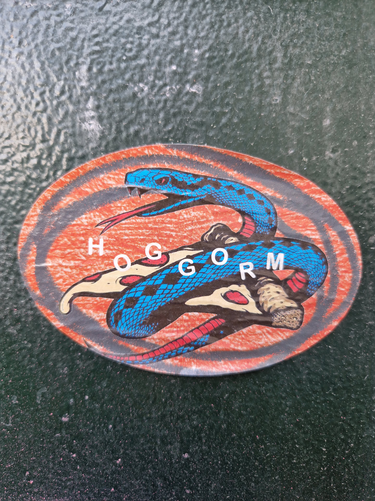 A sticker featuring a blue snake coiled around a pizza. The word "HOGGORM" is written in the center. The style is reminiscent of vintage or retro designs.