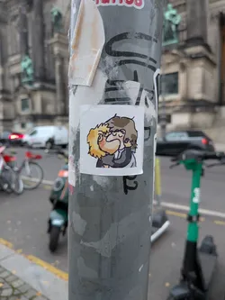 Street sticker A square, white-bordered sticker depicting two cartoon characters in a stylized, chibi-like art style. One character has messy blonde hair and blushed cheeks, while the other has shaggy brownish-grey hair and is cupping the other's cheek while passionately kissing them. Both wear dark clothing, rendered with thick line art and soft cell shading.