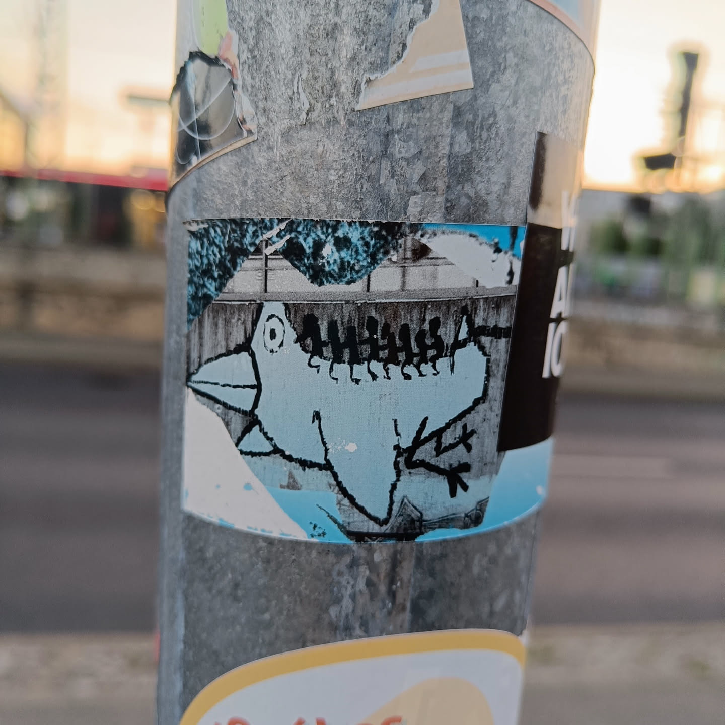 A small sticker featuring a light blue bird illustration.  The bird is stylized and somewhat cartoonish. The background appears to depict a snowy or wintry scene. The sticker is affixed to a metal pole alongside other stickers.