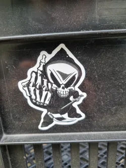 A sticker of a grim reaper giving the middle finger. The sticker is black and white and is on a dark gray surface.