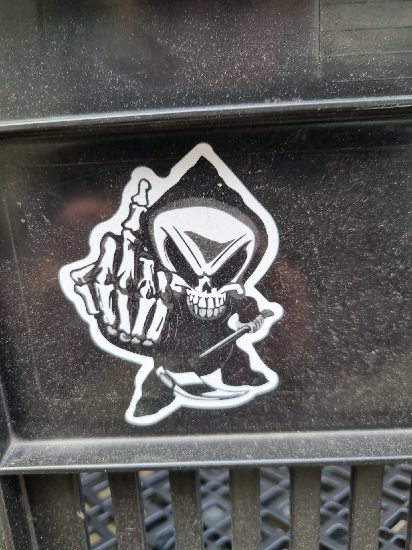 A sticker of a grim reaper giving the middle finger. The sticker is black and white and is on a dark gray surface.