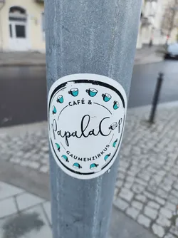 A round sticker advertising Papala Cafe & Gaumenzirkus.  It features a simple design with teal coffee cups surrounding the cafe's name. The sticker is affixed to a metal pole.