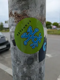 A round green sticker on a metal pole featuring a blue, abstract design in the center. The text "CRUMPLER" is written around the edge of the sticker. The pole shows signs of rust and wear.