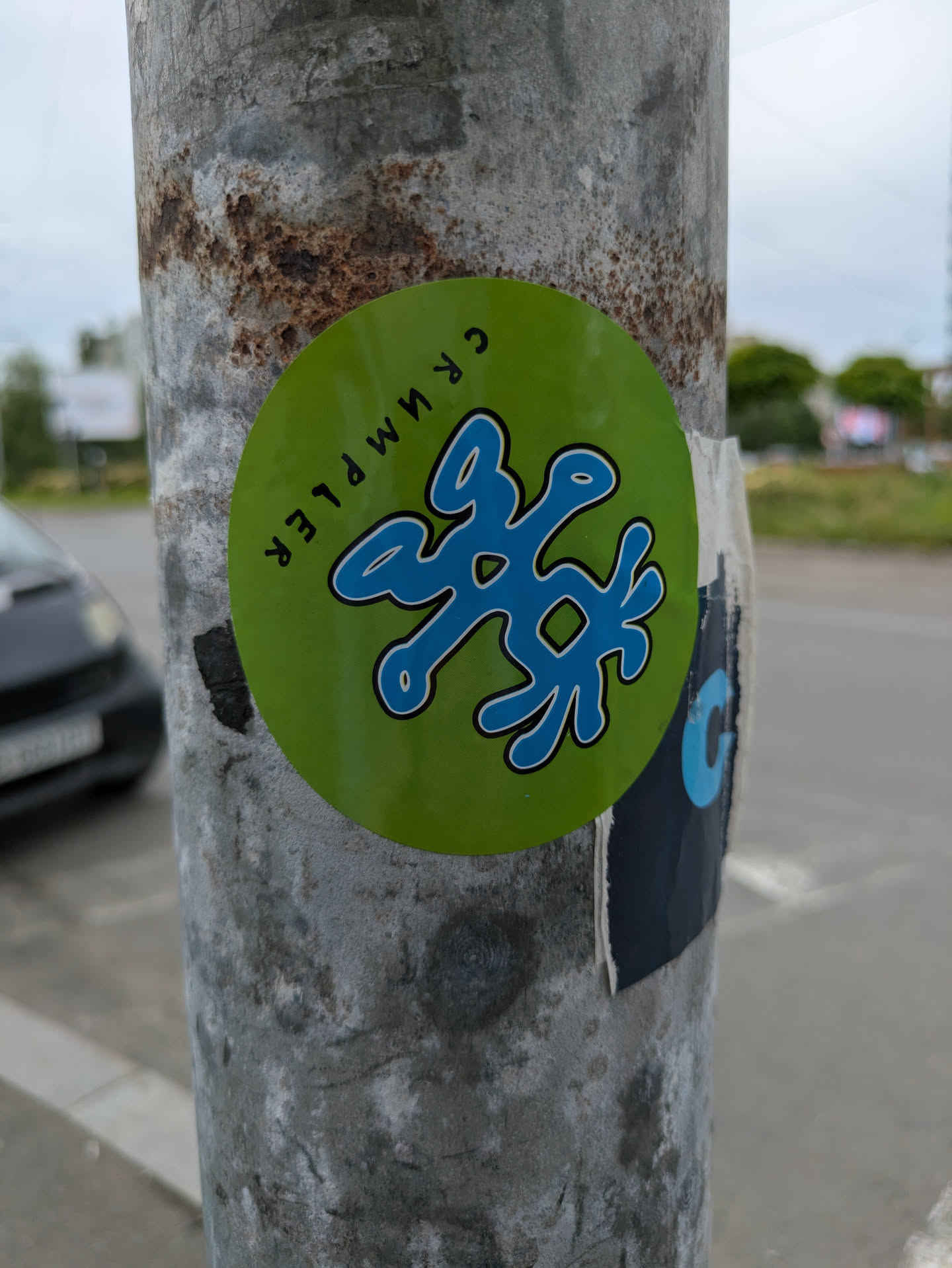 A round green sticker on a metal pole featuring a blue, abstract design in the center. The text "CRUMPLER" is written around the edge of the sticker. The pole shows signs of rust and wear.