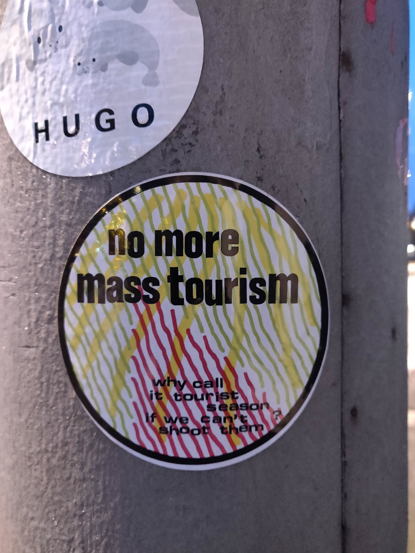 Round sticker with a black outline and a colorful striped pattern. It displays the text "no more mass tourism" in bold black letters. Below it, smaller text reads: "why call it tourist season if we can't shoot them".