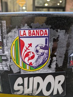 This sticker prominently features the text "LA BANDA" in red letters at the top of a white shield. Below, there's a red fleur-de-lis over a globe with a soccer ball and shoe drawn in purple. The background includes green and yellow bars, and the shield has a purple border.