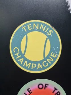A round sticker with a tennis ball graphic in the center and the text "Tennis Champagne" surrounding it. The sticker has a vintage, distressed look, with a teal background and yellow tennis ball.