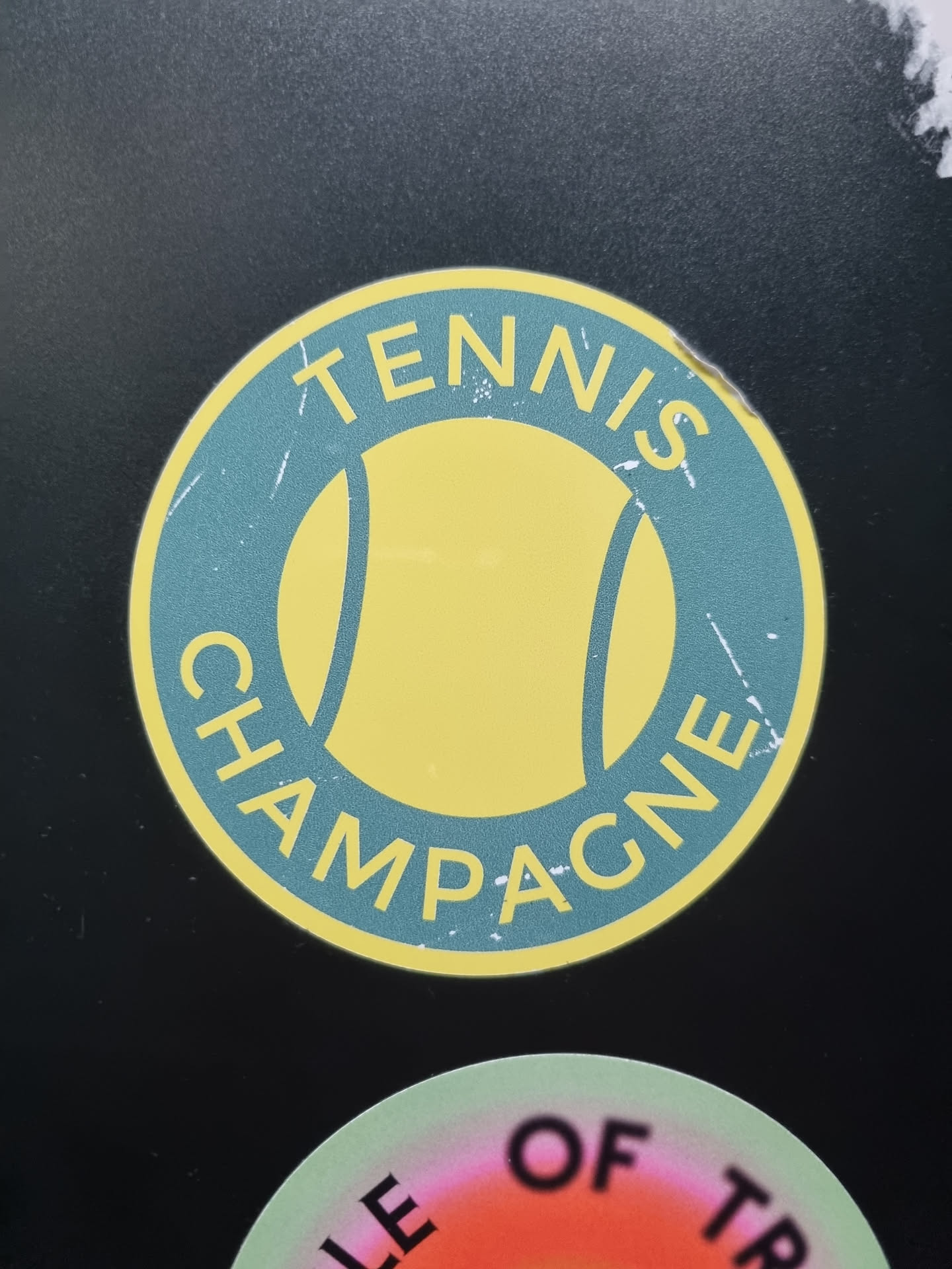 A round sticker with a tennis ball graphic in the center and the text "Tennis Champagne" surrounding it. The sticker has a vintage, distressed look, with a teal background and yellow tennis ball.
