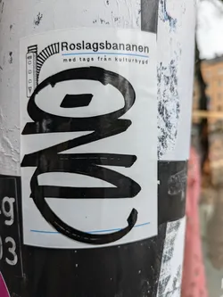 A black and white sticker with a stylized logo and text. The text says Roslagsbanan med tags från kulturbygg.  The logo appears to be an abstract design or a stylized letter or number. The sticker is attached to a pole.