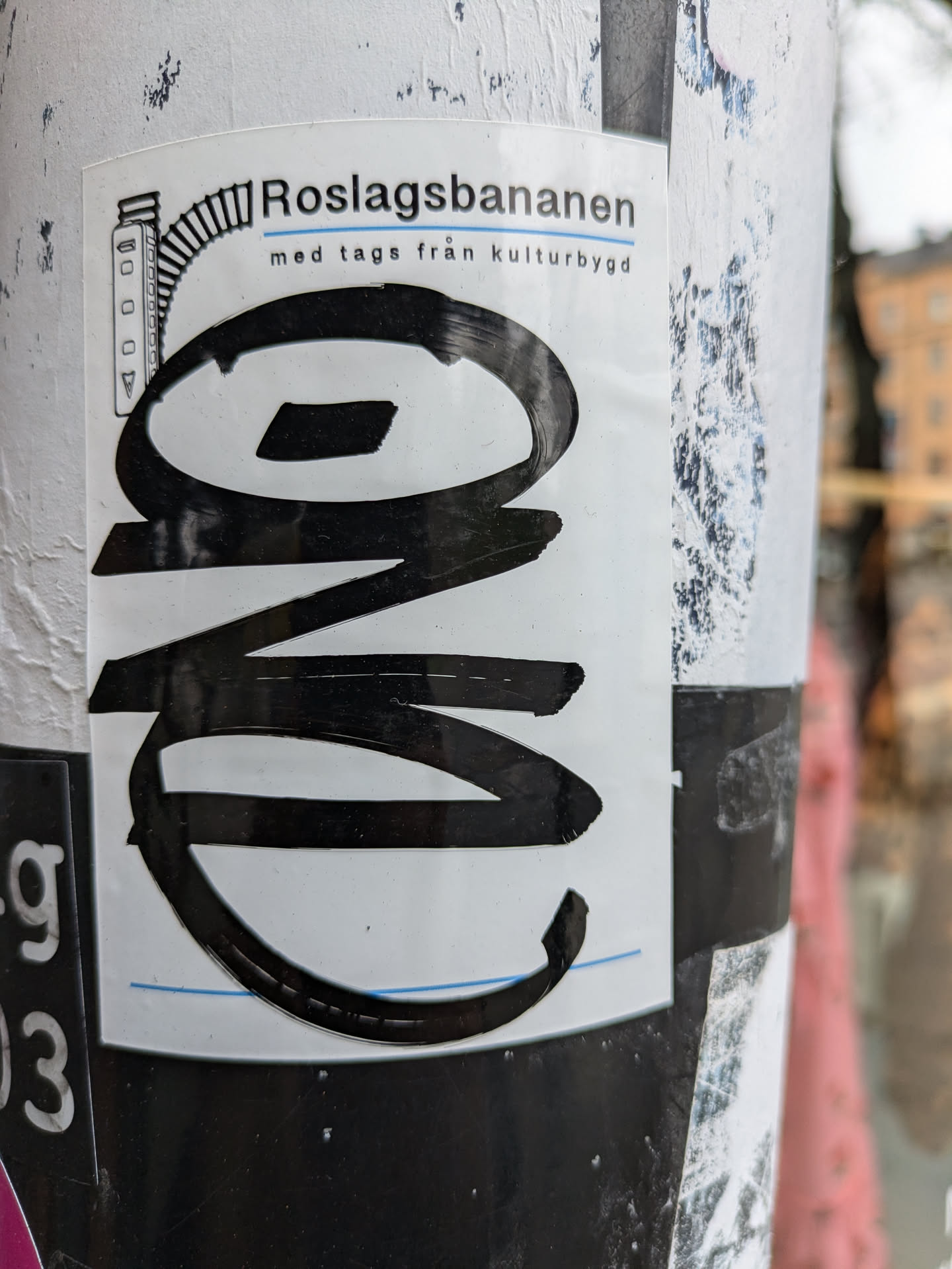 A black and white sticker with a stylized logo and text. The text says Roslagsbanan med tags från kulturbygg.  The logo appears to be an abstract design or a stylized letter or number. The sticker is attached to a pole.