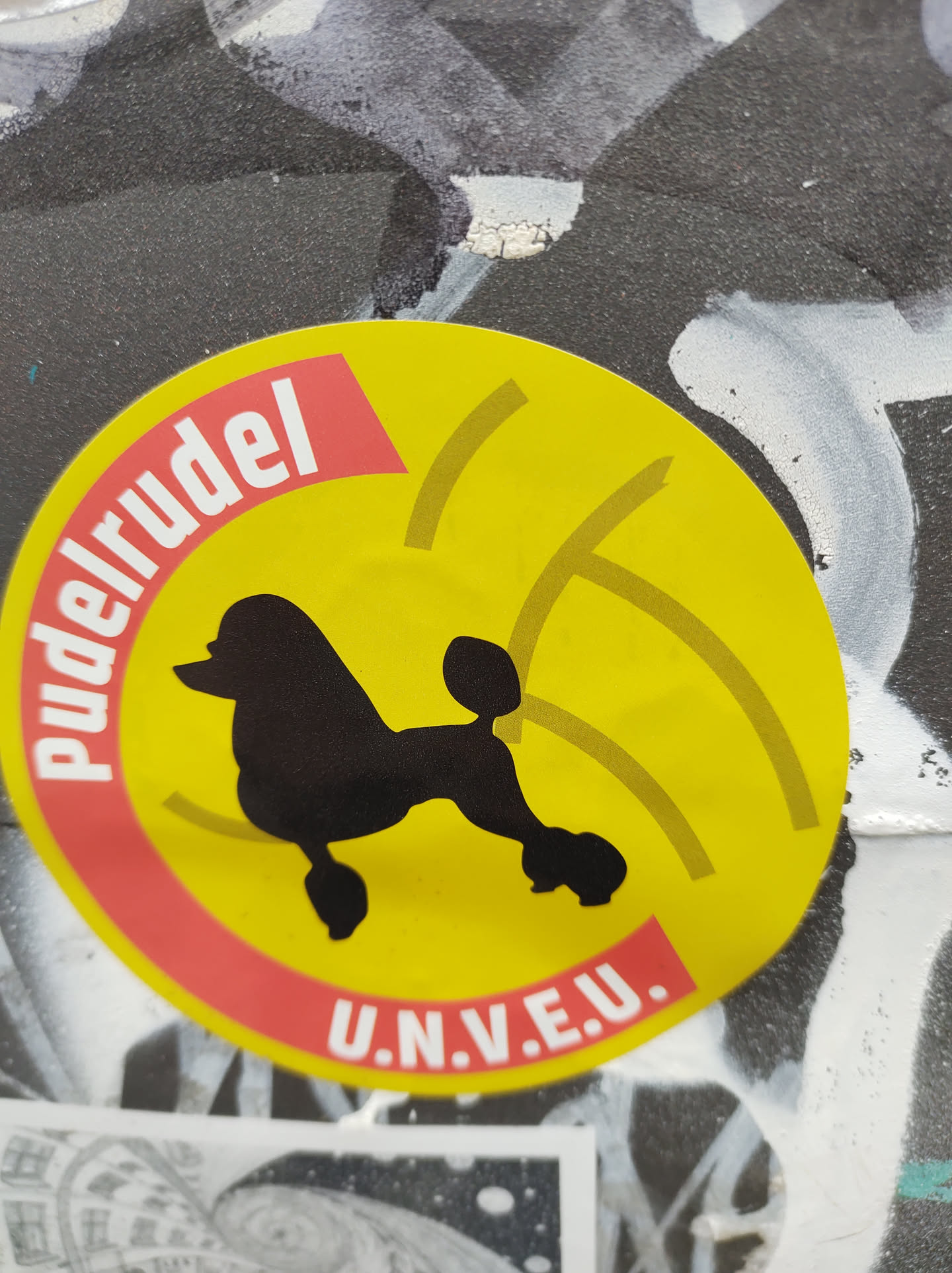 Yellow and red round sticker featuring a silhouette of a poodle and the text "Pudelrudel U.N.V.E.U." The sticker has a design that suggests sound waves around the poodle.