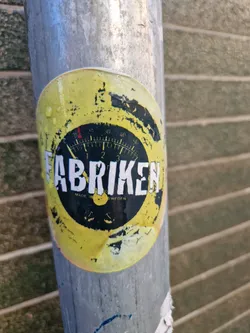 A weathered yellow circular sticker with the word "FABRIKEN" in bold black letters.  A gauge-like graphic is in the center, and the text "MADE IN SWEDEN" is visible below the main text. The sticker shows signs of wear and tear, adding to its character. It is affixed to a metal pole.