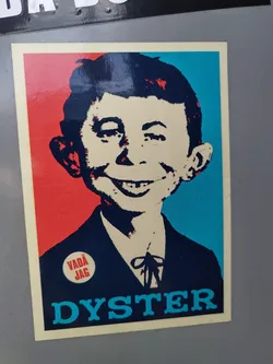 A sticker featuring a stylized portrait of a mischievous-looking boy, reminiscent of Ralph Steadman's illustrations, set against a bold red, white, and blue color scheme. The word "DYSTER" is prominently displayed at the bottom, and a small circular emblem saying "VADA JAG" is present on his chest.