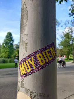 A yellow sticker with the words "MUY BIEN" (Spanish for "very good") on a textured, red, white, and blue background. The sticker is affixed to a weathered grey pole. 