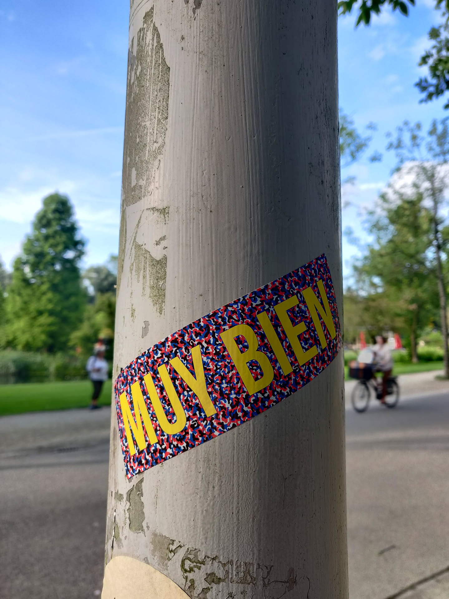 A yellow sticker with the words "MUY BIEN" (Spanish for "very good") on a textured, red, white, and blue background. The sticker is affixed to a weathered grey pole. 