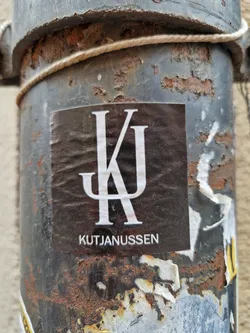 A black sticker with a white monogram of KJ and the text Kutjanussen below. The sticker is affixed to a rusty metal pipe.