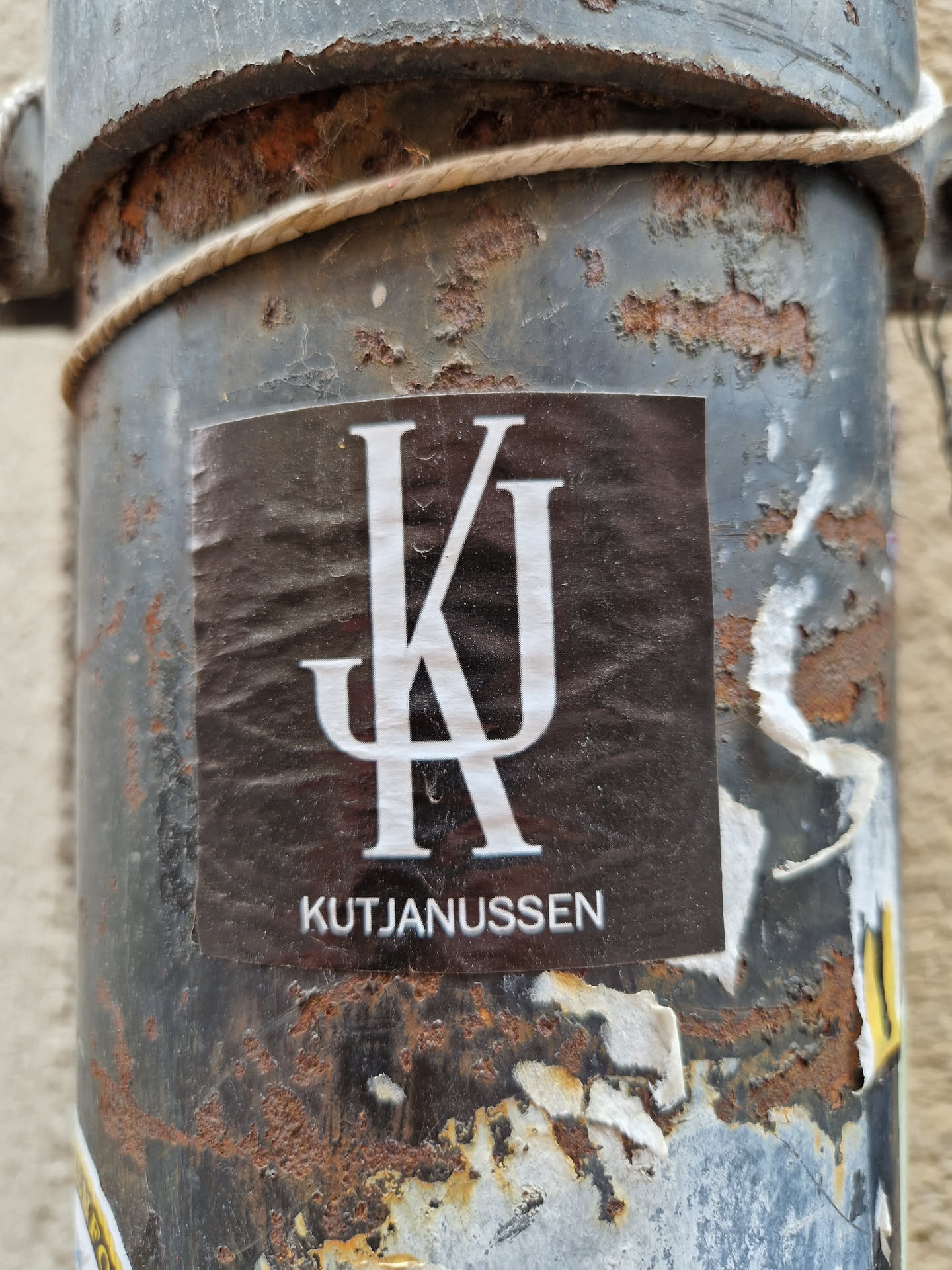 A black sticker with a white monogram of KJ and the text Kutjanussen below. The sticker is affixed to a rusty metal pipe.