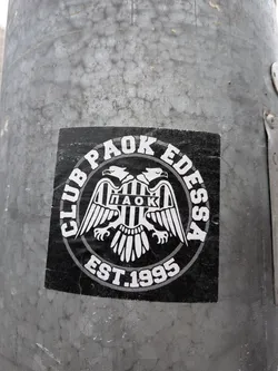 A circular, black and white sticker featuring a stylized double-headed eagle as its central motif. The eagle has outstretched wings and holds a shield on its chest, which displays vertical black and white stripes and the Greek letters 'ΠΑΟΚ' (PAOK). Encircling the eagle, in an arc at the top, is the text 'CLUB PAOK EDESSA' in white capital letters. Below the eagle, in a similar arc, is 'EST.1995'. The entire design is bordered by a white and grey double ring. The sticker is applied to a textured grey metal pole and shows minor signs of wear and creasing.